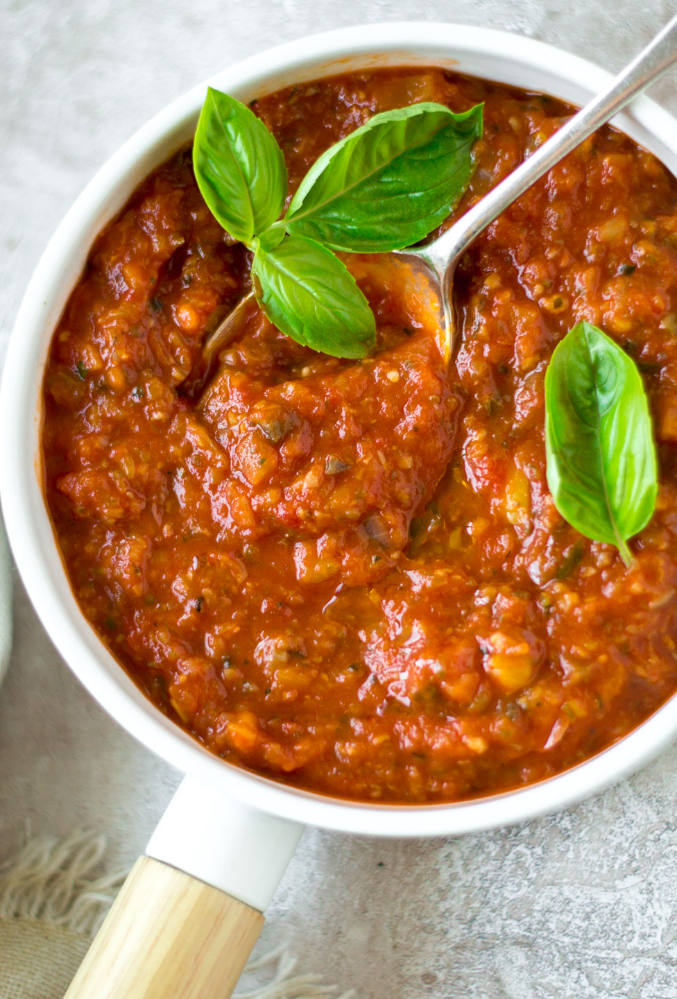 Top down view marinara sauce in a white dish with spoon and basil leaves