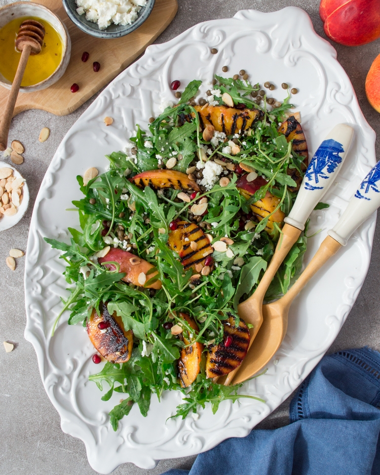 A white plate with grilled nectarines, arugula, lentils & salad serving spoons 