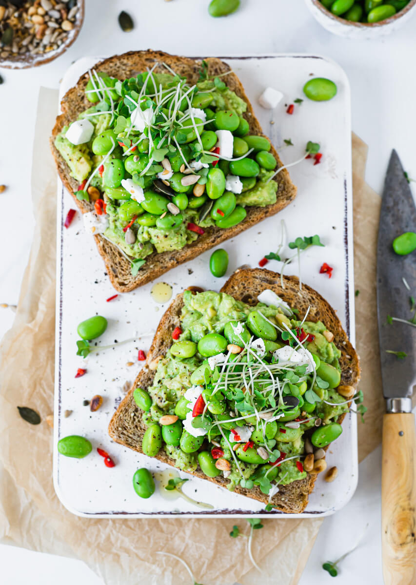 Overhead view of avocado toast with edamame beans & feta on a white board  