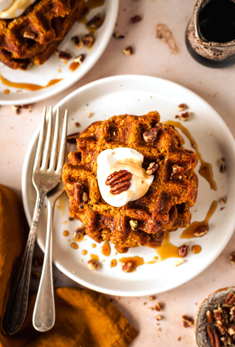 Overhead view of pumpkin waffles on a white plate topped with pecans, yoghurt and maple syrup  