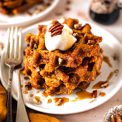 Pumpkin waffles on a white plate topped with pecans, yoghurt and maple syrup